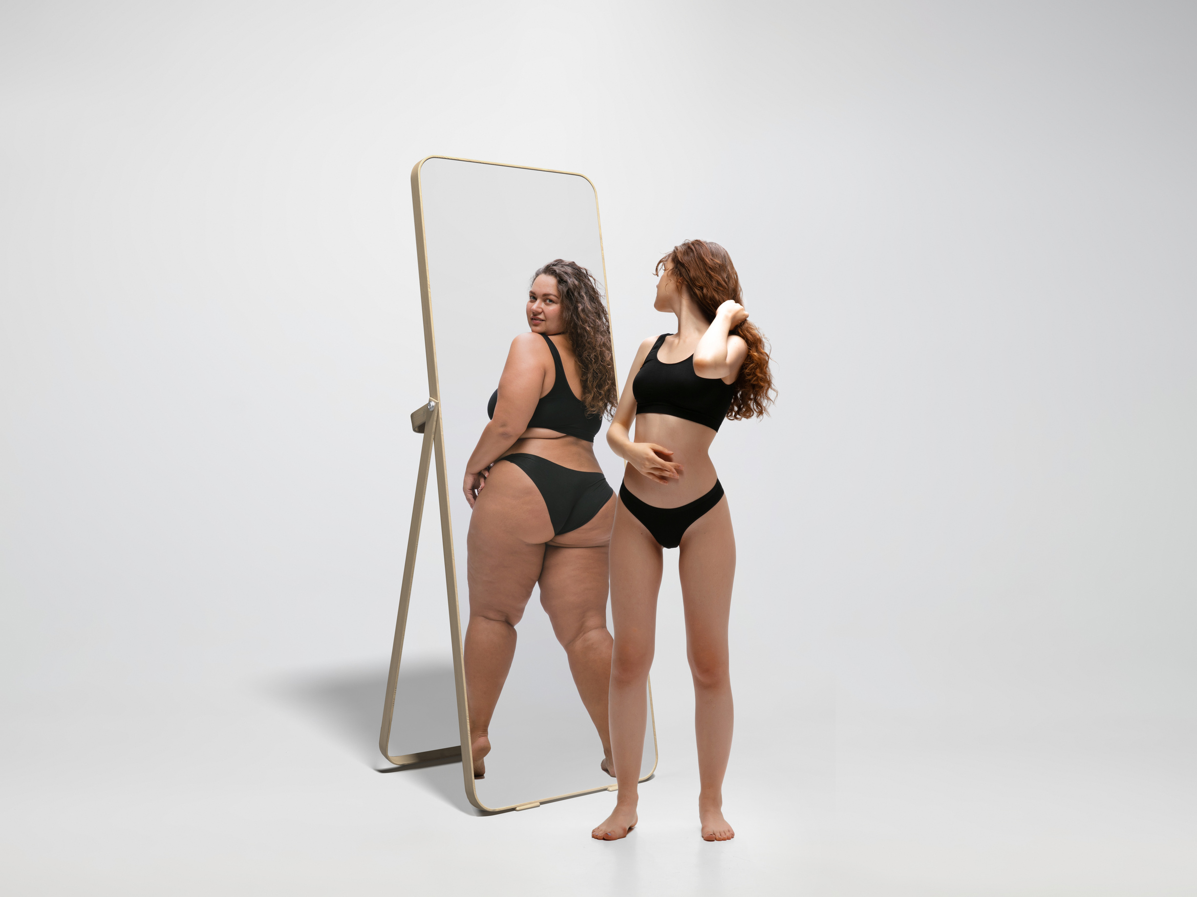 Young fit, slim woman looking at fat girl in mirror's reflection on white background